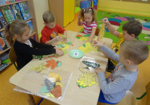 Pięcioro dzieci siedzi przy stole na którym rozłożone są kolorowe liście papierowe oraz owoce. Dzieci dobierają owoce do liści drzew.